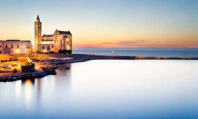 Tramonto sul porto di Trani, con la cattedrale illuminata che domina il lungomare. I vicoli del centro storico di Trani si intravedono tra gli edifici.