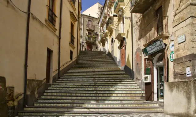 Una scalinata decorata con maioliche colorate sale tra edifici storici. Un negozio di fiori e souvenir invita a scoprire cosa vedere a Vizzini.