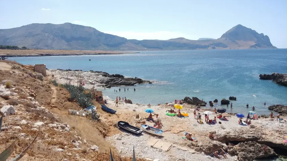 La **spiaggia di Isulidda** è affollata di bagnanti sotto ombrelloni colorati. Il mare azzurro invita a un bagno rinfrescante, mentre le montagne sullo sfondo creano un panorama mozzafiato.