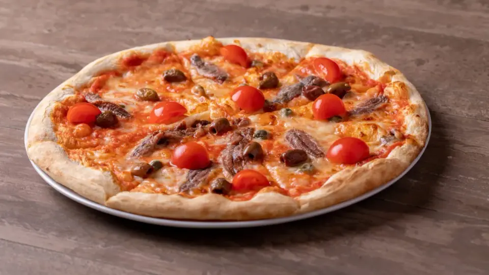 Pizza fuata siciliana con pomodorini, acciughe, olive e capperi su un piatto bianco, su un tavolo di legno.