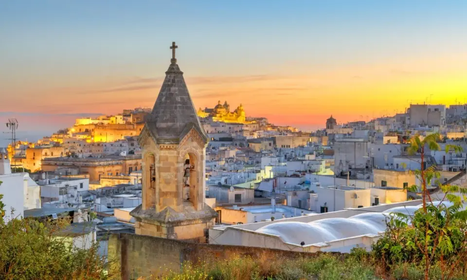 Campanile in primo piano, sullo sfondo la città bianca di Ostuni illuminata dal tramonto. Perfetto per scoprire cosa vedere a Ostuni in mezza giornata.