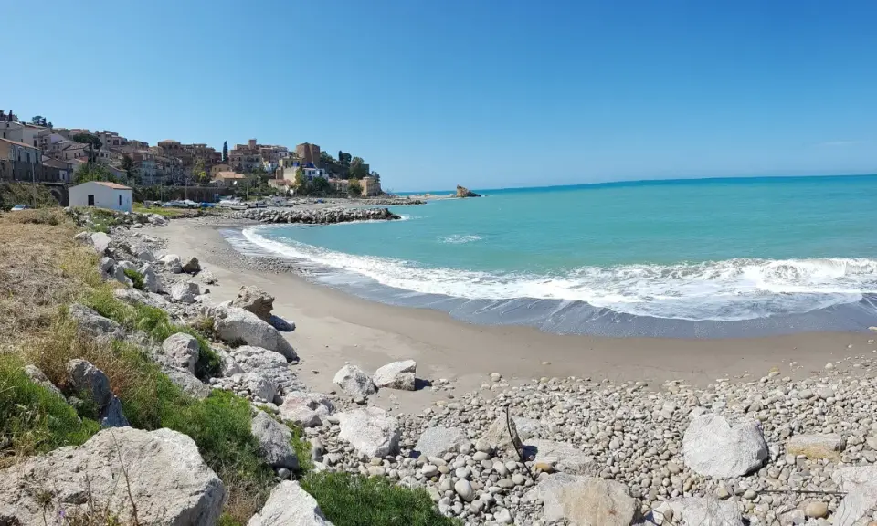Spiaggia di Lampare a Tusa, con mare turchese, case colorate sul promontorio e rocce sulla riva.