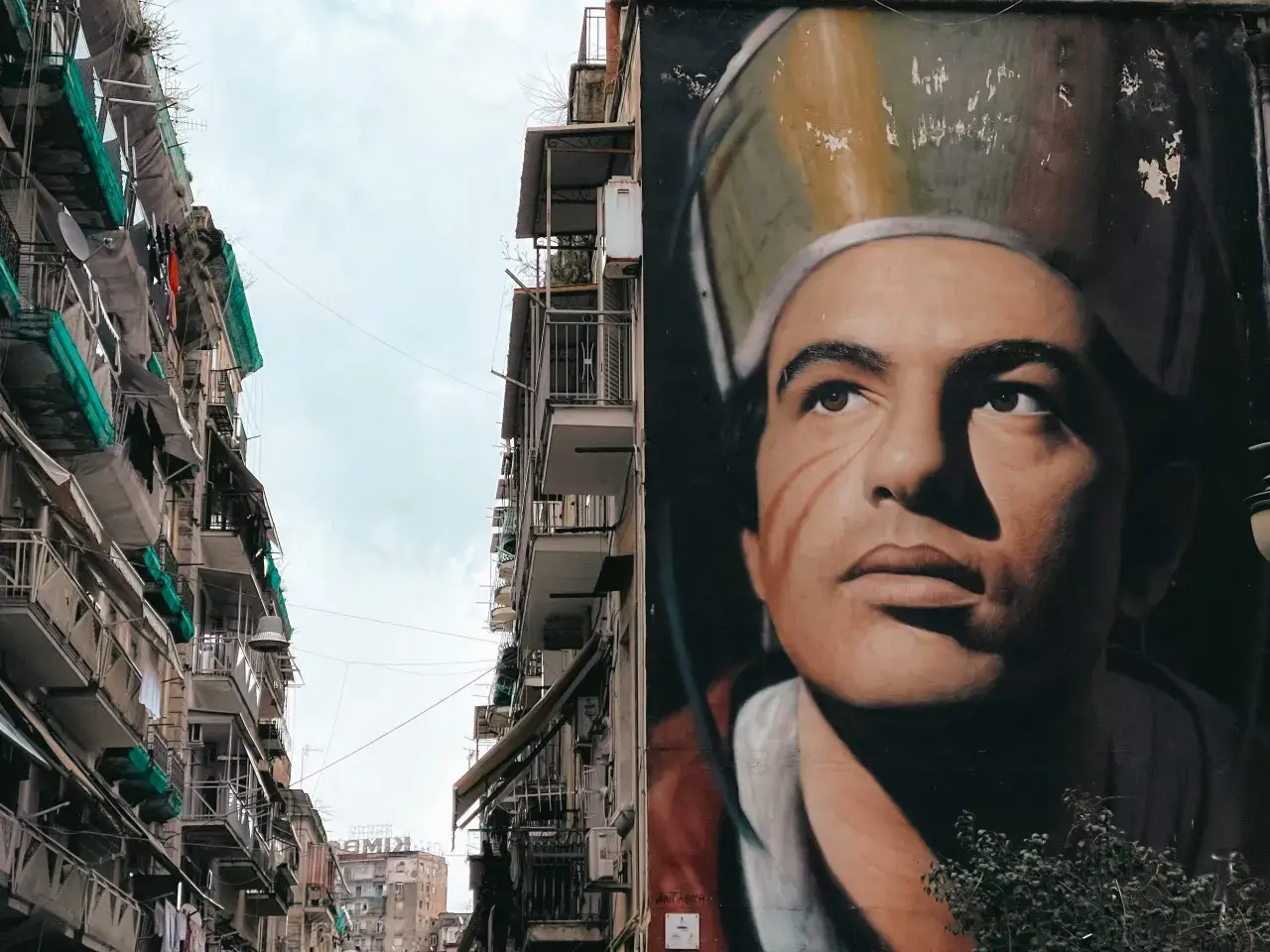 Un imponente murales napoli raffigura un volto intenso, con un cappello particolare. Accanto, palazzi con balconi e panni stesi.