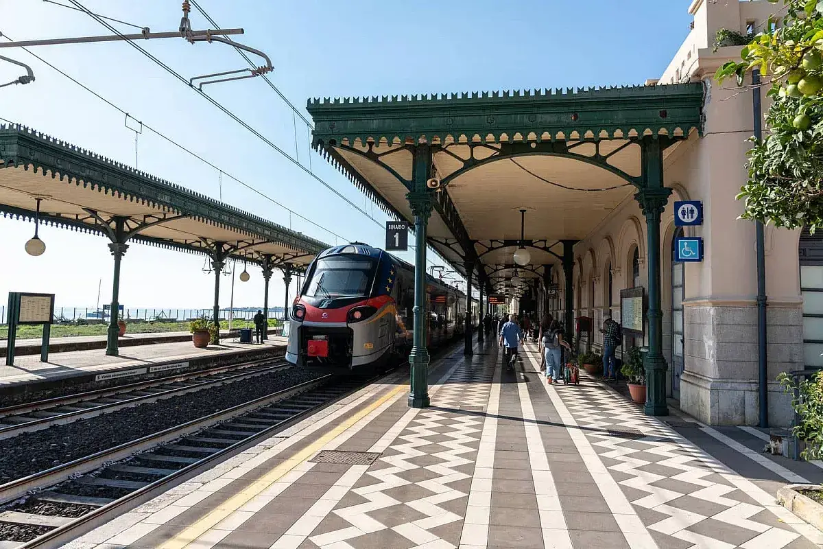 Treno in arrivo alla **stazione Taormina** sotto un portico verde. Persone attendono sul binario 1.