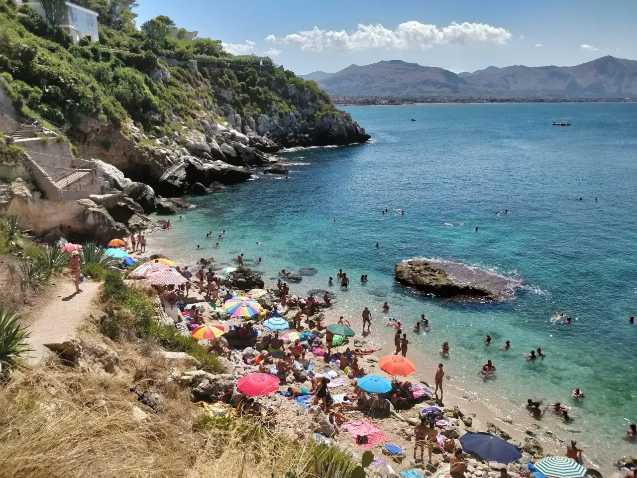 La Baia dei Francesi è affollata di bagnanti sotto ombrelloni colorati, con acque cristalline e montagne sullo sfondo.