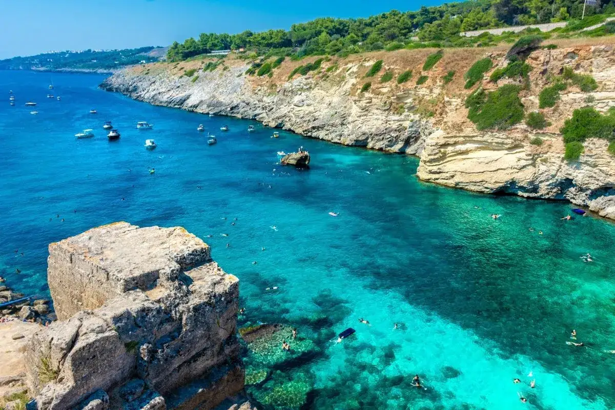 Scogliera a picco sul mare cristallino, con barche e bagnanti che godono della spiaggia 100 gradini Mongerbino.