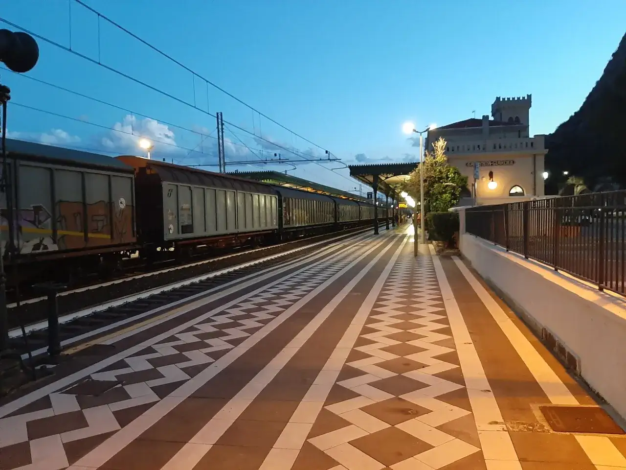 Stazione Taormina: un treno merci attende sul binario, illuminato da luci soffuse. Il cielo azzurro della sera fa da sfondo.