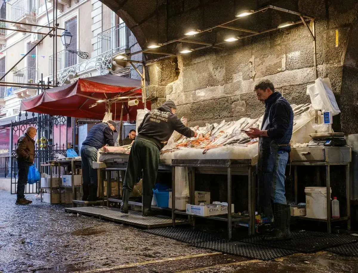 Mercato a Catania tutti i giorni: pescivendoli al lavoro sotto un arco, con pesce fresco esposto su un banco ghiacciato.