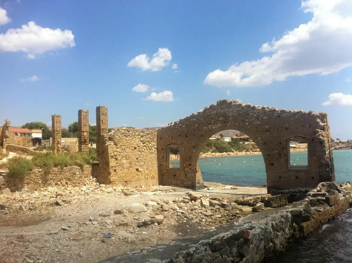 Rovine della tonnara di Avola, con un arco in pietra che si affaccia su una spiaggia affollata e il mare azzurro.