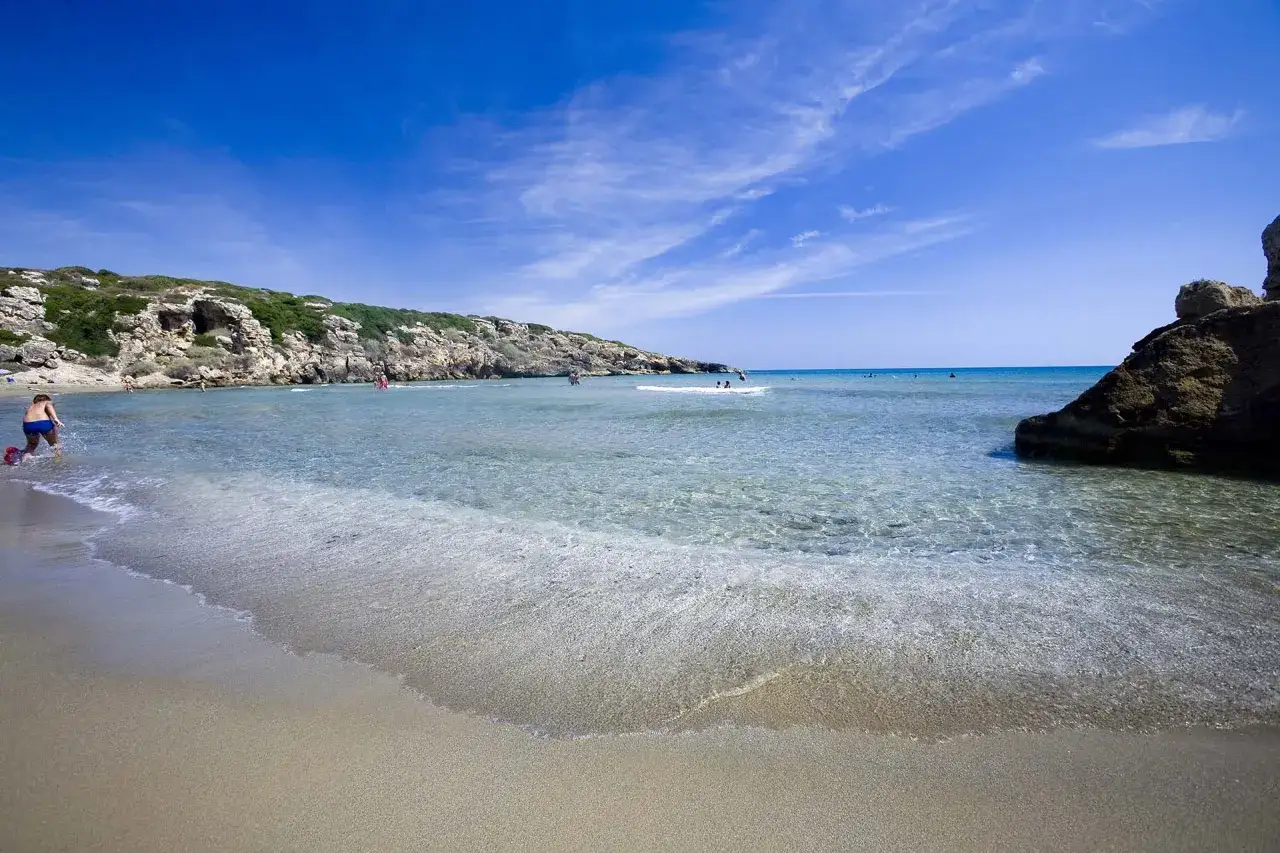 Bambino gioca sulla sabbia dorata di Calamosche, Sicilia, con acque cristalline e rocce a picco sul mare.