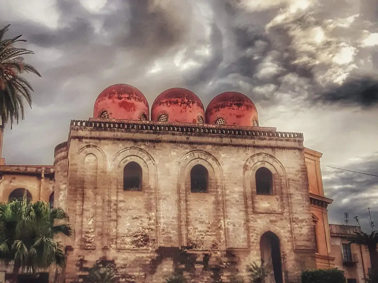 Le iconiche cupole rosse di Palermo si stagliano contro un cielo tempestoso, con palme che incorniciano l'antica architettura.