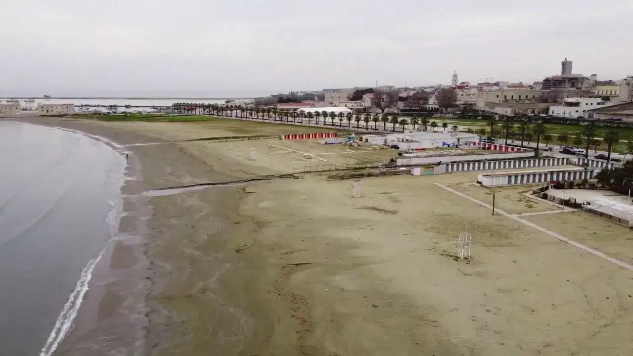 La spiaggia Baia delle Sirene a Trani, con le cabine, la passeggiata alberata e il mare calmo sotto un cielo nuvoloso.