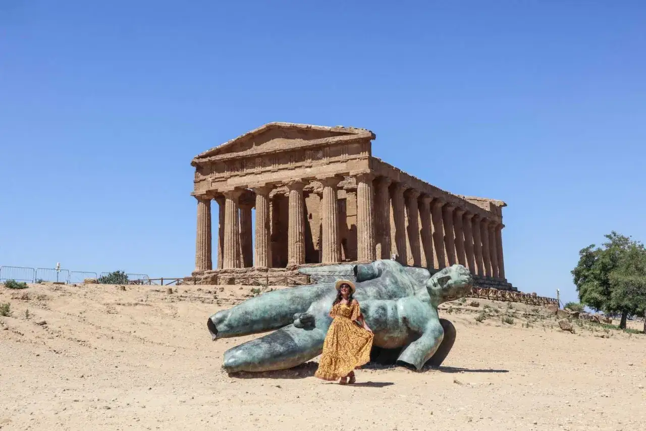 Donna con abito giallo posa accanto a una scultura di un gigante, con il Tempio della Concordia sullo sfondo. Il significato di Akragas &egrave; qui tangibile.