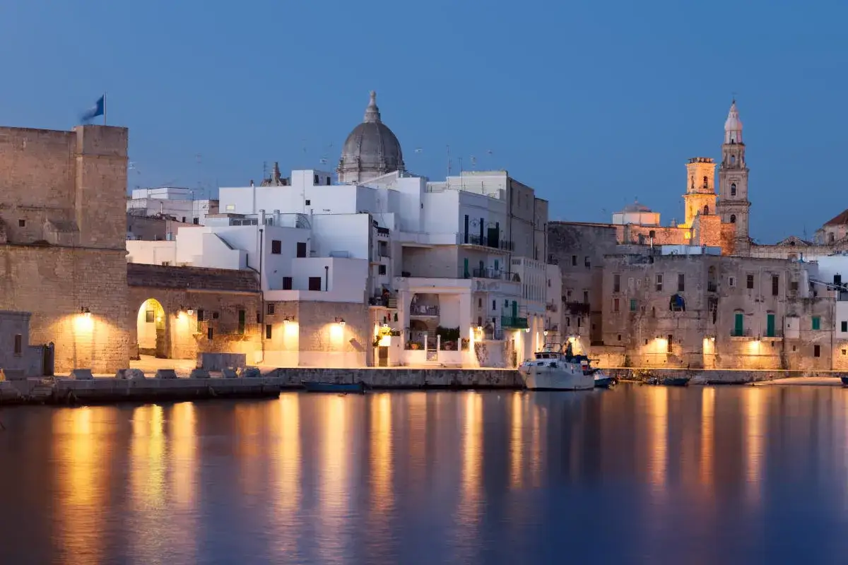 Monopoli di sera: un borgo antico illuminato, con il porto e le chiese che si specchiano sull'acqua.