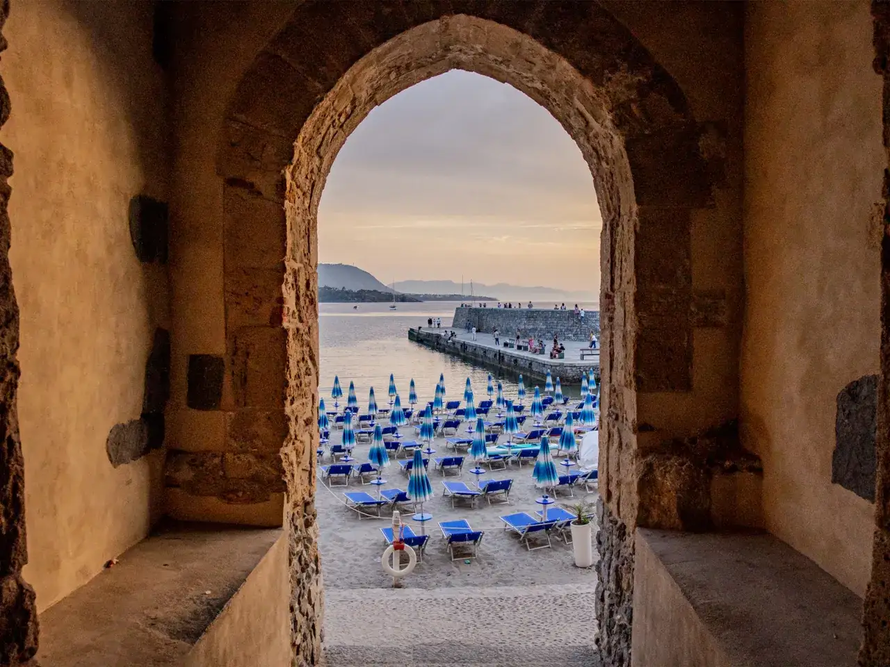 Arco antico incornicia la spiaggia di Cefal&ugrave; con ombrelloni blu e lettini, vista sul mare e sul molo affollato.