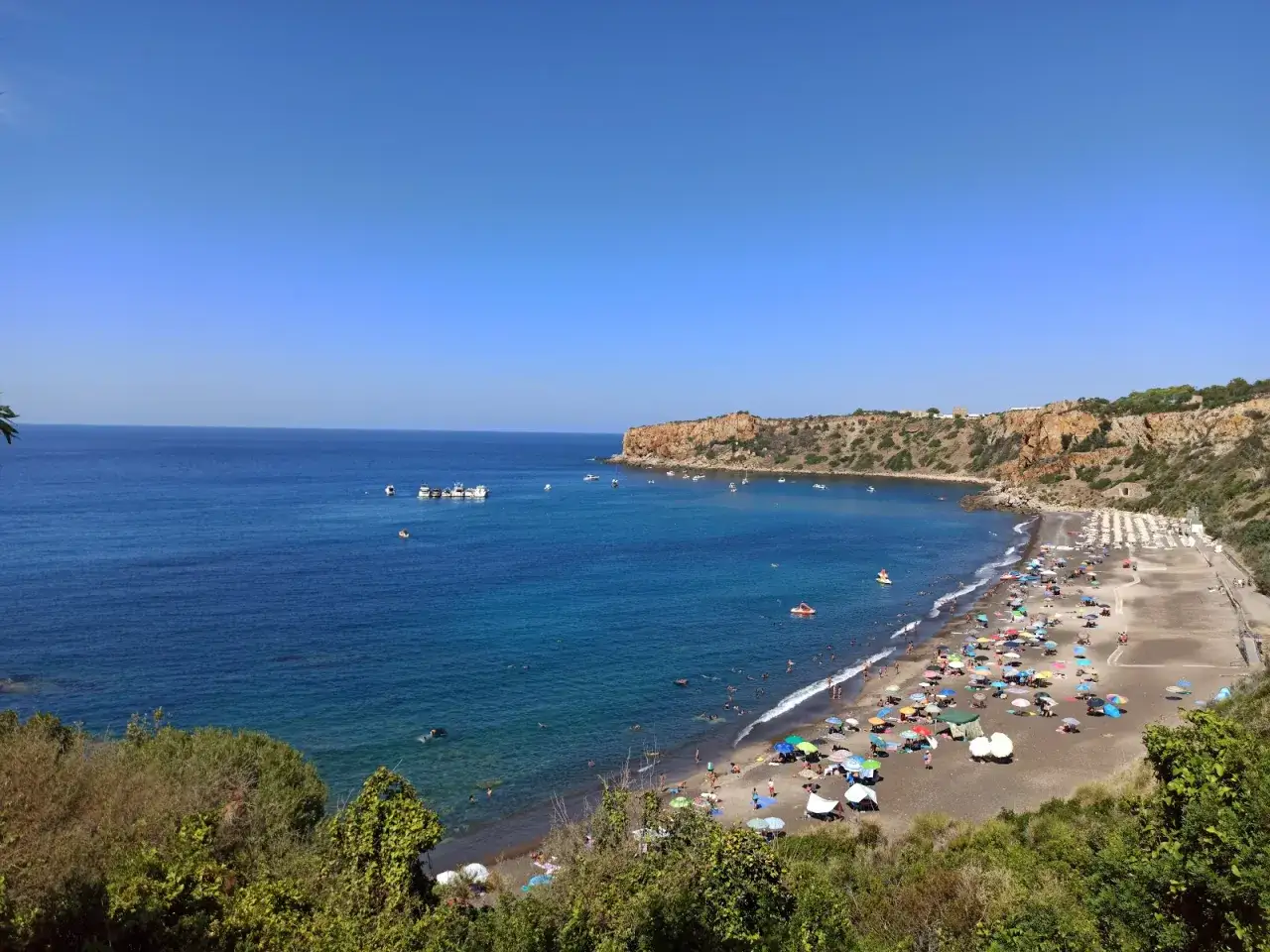 Spiaggia affollata con ombrelloni colorati e gente che nuota, con la costa rocciosa e la torre conca sullo sfondo.