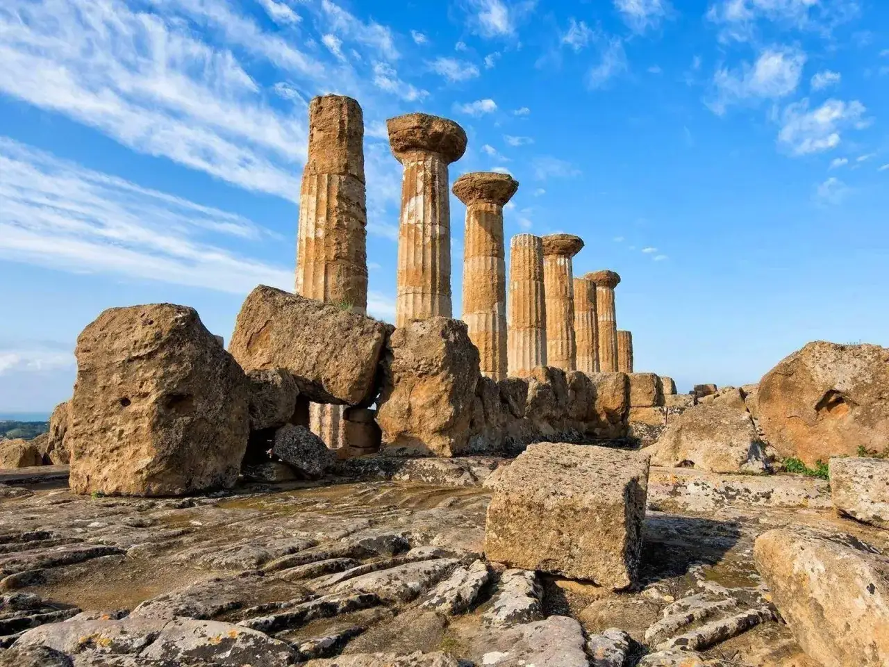 Colonne doriche imponenti si ergono tra rocce antiche, testimoni della Valle dei Templi storia. Cielo azzurro con nuvole bianche.