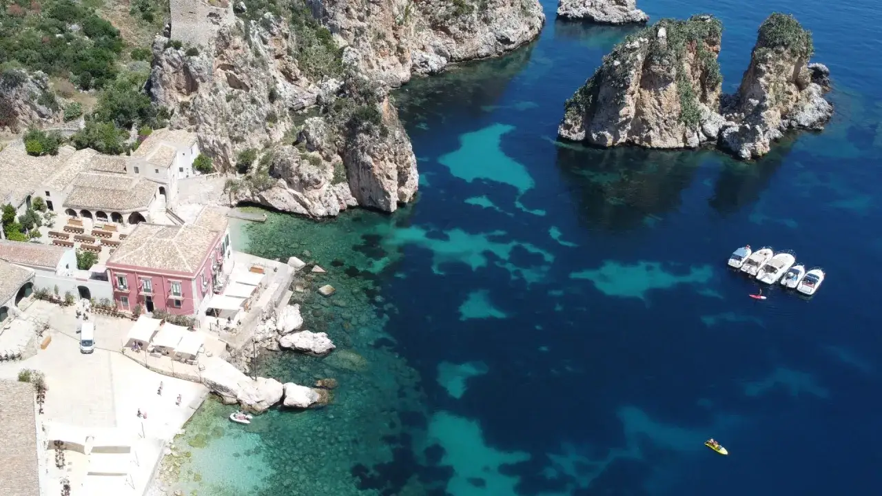 Vista aerea della costa siciliana, con acque cristalline e scogliere. Un borgo con edifici storici e un rosa acceso, ideale per capire come arrivare a Scopello.