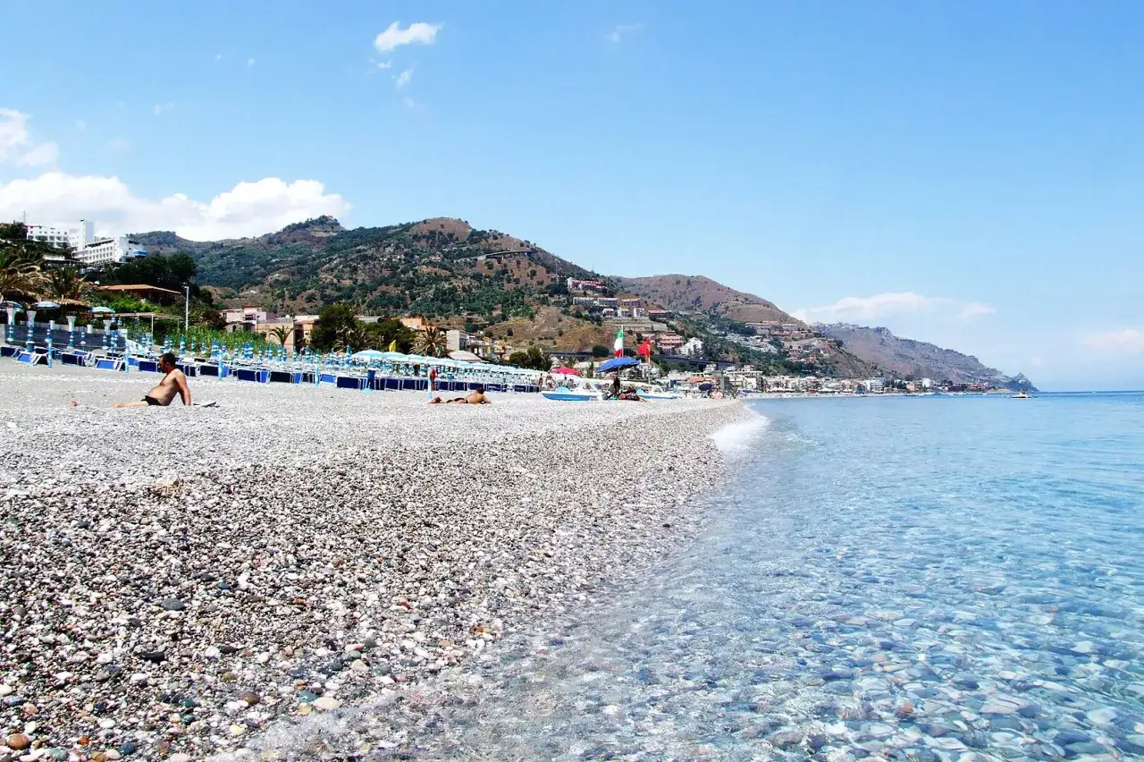 Sole sulla spiaggia di Mazzeo, con il mare cristallino che lambisce la riva di ciottoli e le colline verdi sullo sfondo.