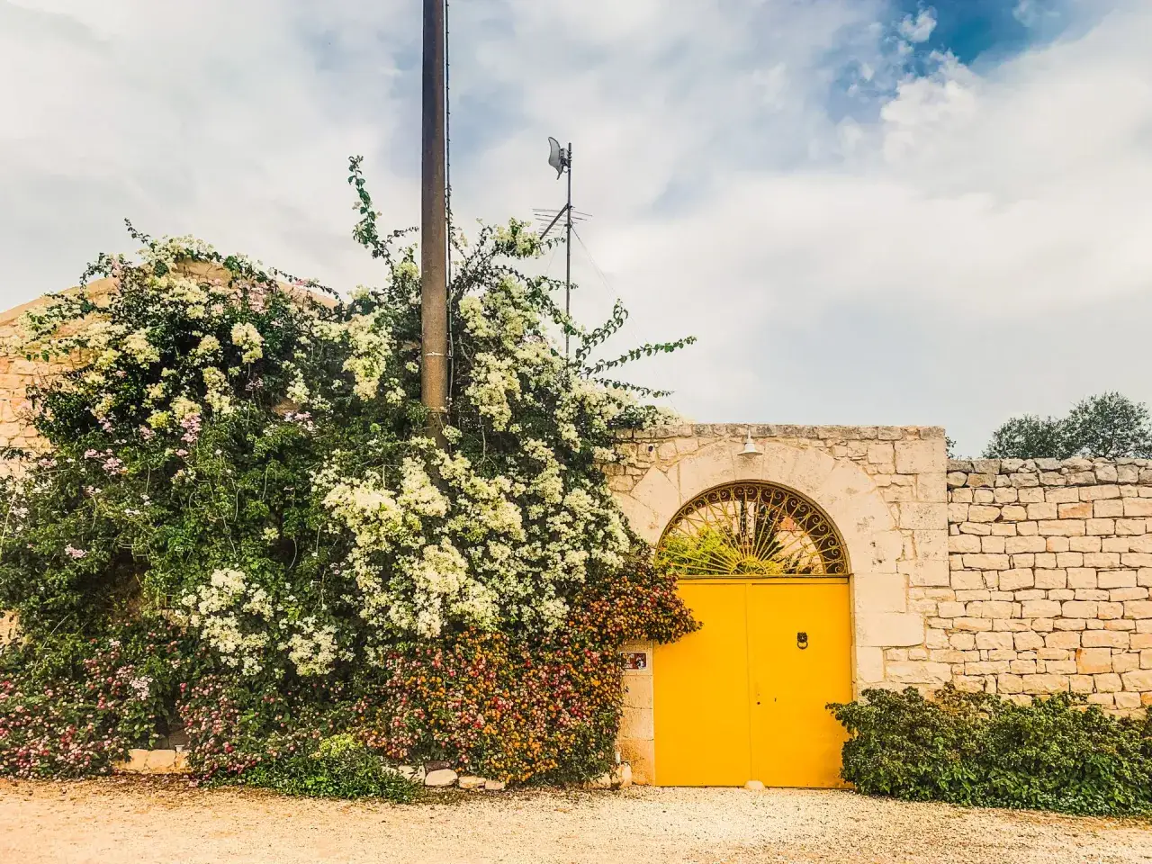 Un cancello giallo brillante si apre su un cortile fiorito. Un muro di pietra circonda il baglio, cos'è? Un luogo di pace e bellezza.