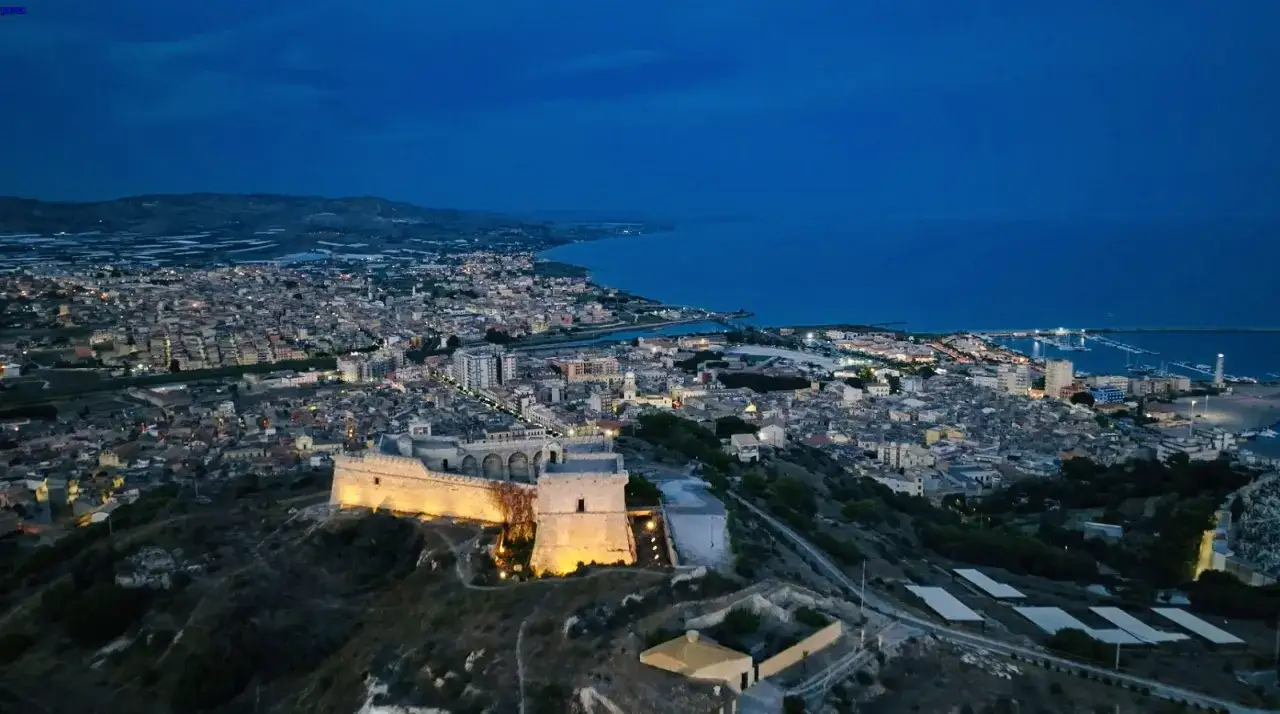 La rocca licata domina la citt&agrave; illuminata al crepuscolo, con il mare blu sullo sfondo.