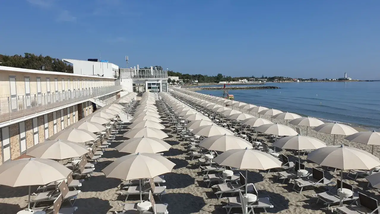 File di ombrelloni aperti sulla sabbia dorata del lido verde San Cataldo, con il mare azzurro e un faro in lontananza.