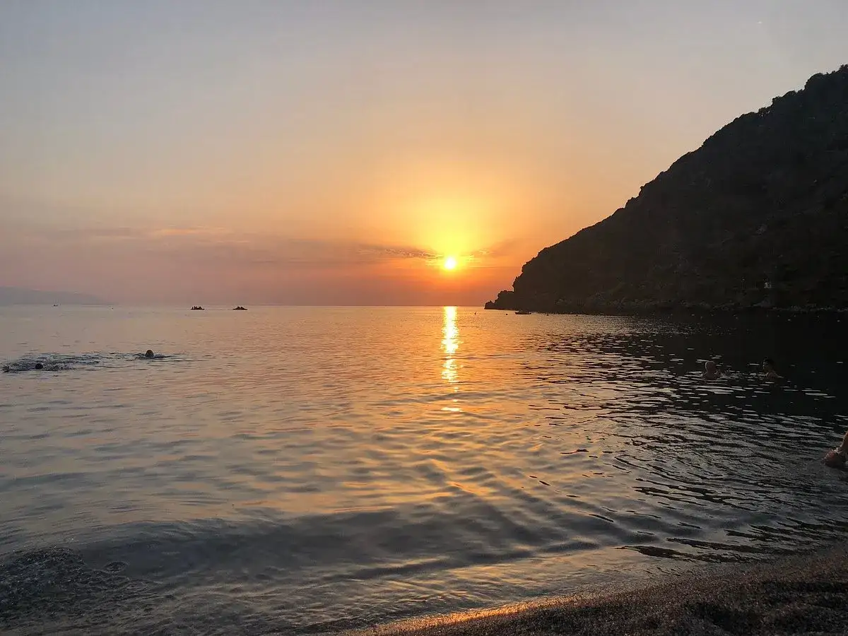 Tramonto dorato sulle spiagge di Milazzo, con bagnanti che si godono le ultime luci del giorno in un mare calmo.