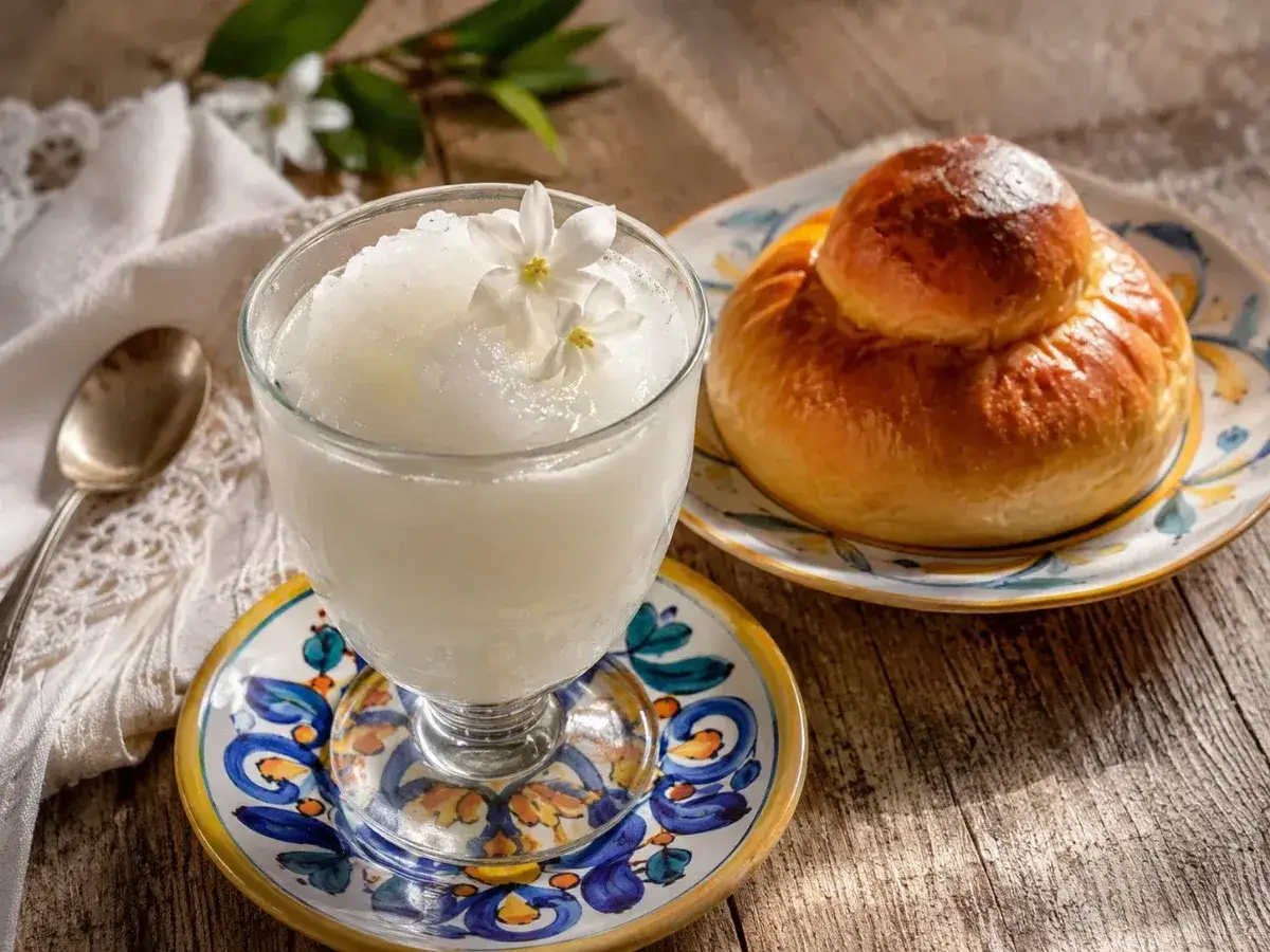 Rinfrescante granita al gelsomino, guarnita con fiori, accanto a una brioche su un piatto decorato. Un cucchiaino d'argento completa la scena.