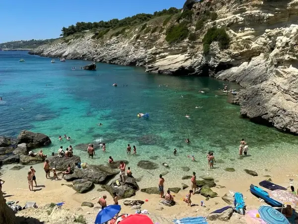 Spiaggia 100 gradini Mongerbino, con acque cristalline e scogliere rocciose. Persone nuotano e prendono il sole in una giornata estiva.