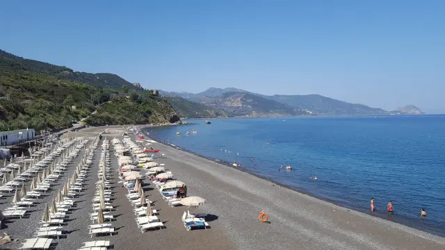 Spiaggia Torre Conca: file di lettini e ombrelloni su ghiaia scura, mare azzurro e colline verdi sullo sfondo.