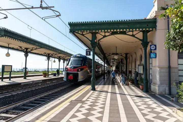 Treno in arrivo alla **stazione Taormina** sotto un portico verde. Persone attendono sul binario 1.