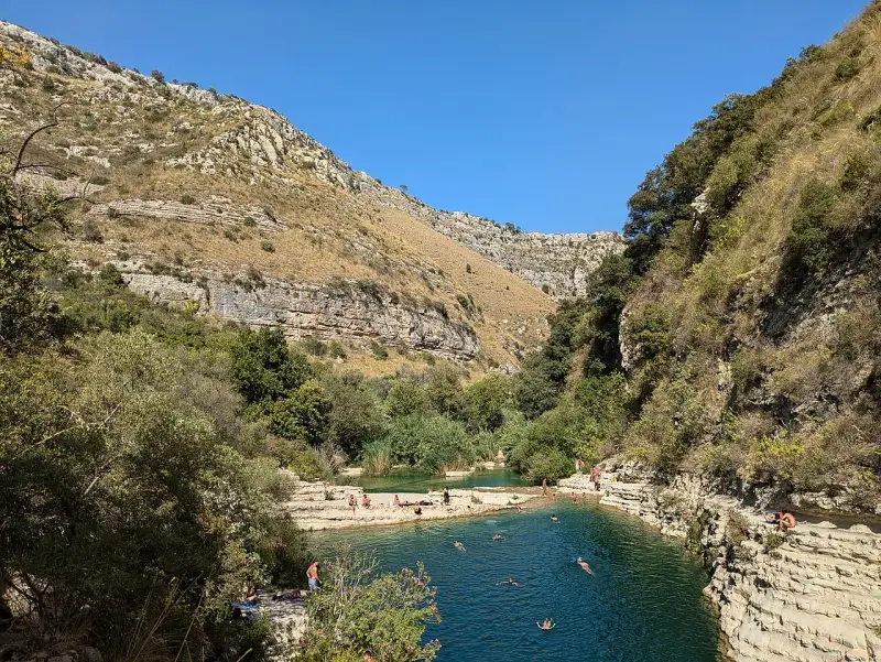 Gente si gode il sole e nuota nei laghetti d'Avola, circondati da rocce e vegetazione lussureggiante sotto un cielo azzurro.