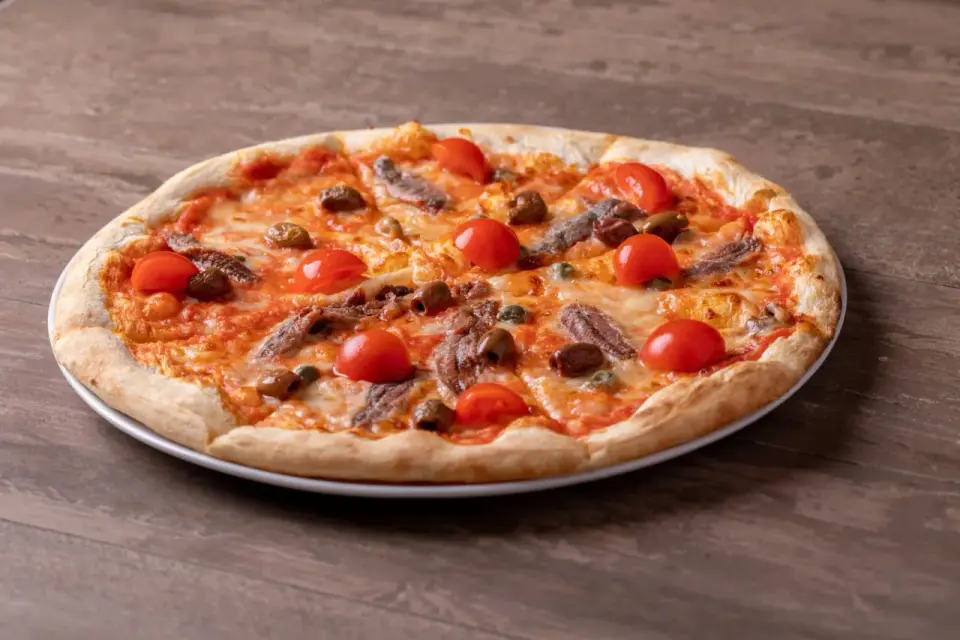 Pizza fuata siciliana con pomodorini, acciughe, olive e capperi su un piatto bianco, su un tavolo di legno.